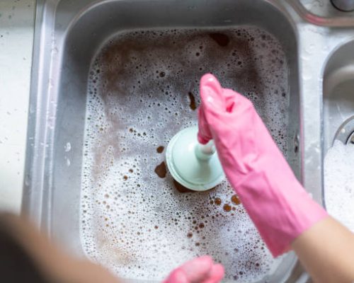 Woman Using Plunger In Blocked Kitchen Sink To Unclog Drain