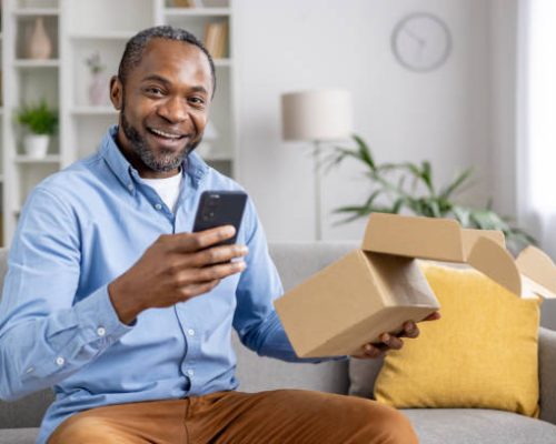Portrait of an African-American man sitting on the sofa at home, holding an open parcel box in his hand, using a mobile phone, looking and smiling at the camera.