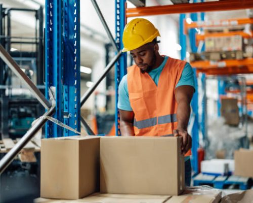 African american warehouse worker is seen handling boxes in a large storage area, equipped with shelves and racks, the worker is dressed in a hard hat and safety vest, ensuring orderliness.
